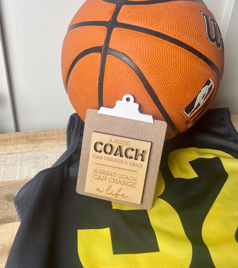 May include: A wooden sign with a clipboard design that reads "A good coach can change a game. A great coach can change a life." The sign is sitting on top of a black and yellow basketball jersey and a basketball.