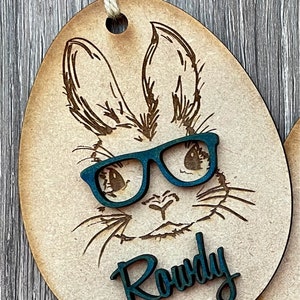 May include: A wooden Easter egg ornament with a laser-cut design of a bunny wearing blue glasses. The bunny's name, "Rowdy", is written in blue lettering below the bunny's face.