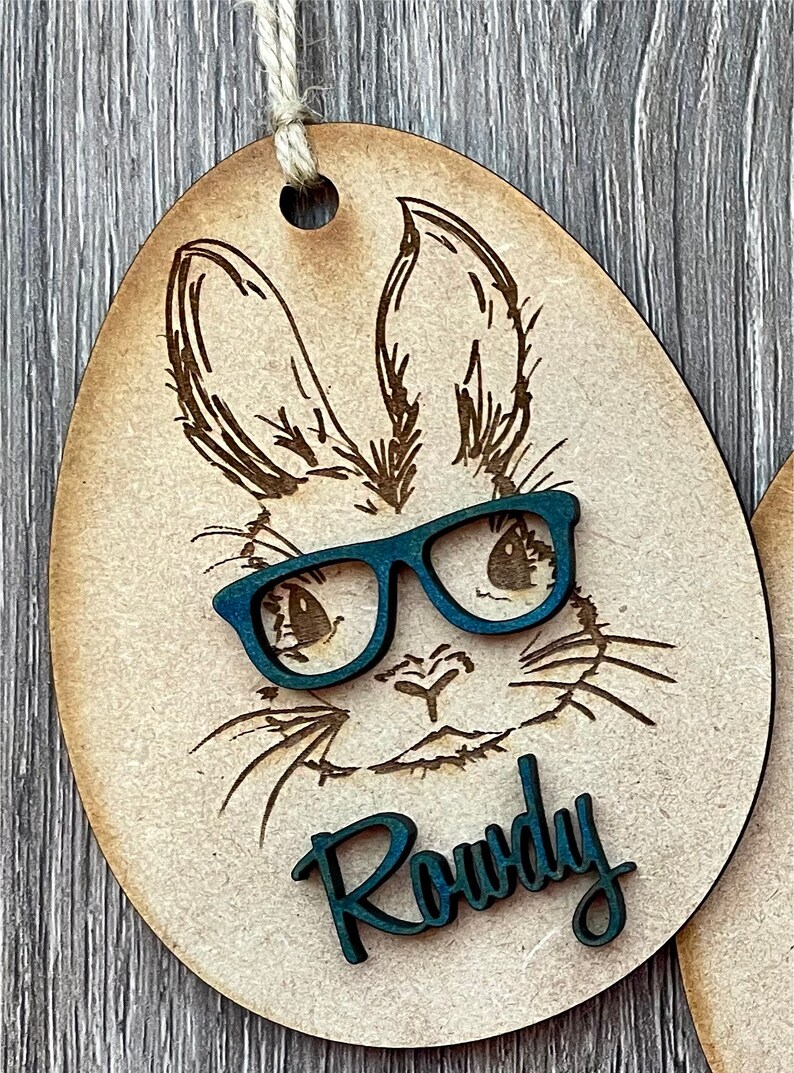 May include: A wooden Easter egg ornament with a laser-cut design of a bunny wearing blue glasses. The bunny's name, "Rowdy", is written in blue lettering below the bunny's face.