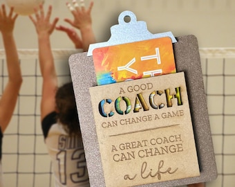 May include: A clipboard-shaped gift card holder with a silver top and a brown, glittery base. The holder has a slot for a gift card and a cutout design that reads "A GOOD COACH CAN CHANGE A GAME, A GREAT COACH CAN CHANGE A LIFE."