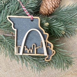 May include: Wooden Christmas ornament in the shape of the state of Missouri, with a glittery gray interior and a cutout of the Gateway Arch and city skyline. The ornament has a red and white string for hanging.