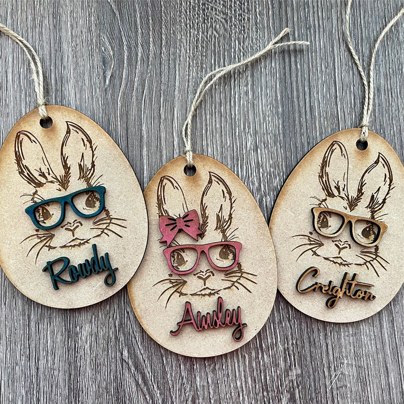 May include: Three wooden Easter egg ornaments with bunny faces, glasses, and bows. The names Rowdy, Ainsley, and Creighton are written on the ornaments.