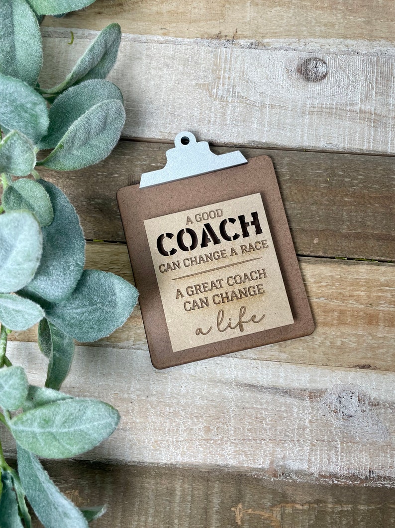 May include: A wooden clipboard with a brown background and white text that reads "A good coach can change a race. A great coach can change a life."