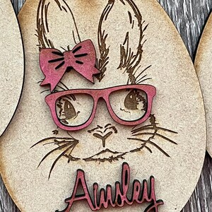 May include: Wooden Easter egg ornament with a laser-cut design of a bunny wearing pink glasses and a pink bow. The name "Ainsley" is written in pink on the ornament.