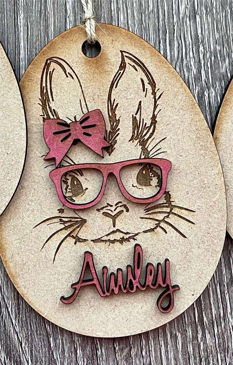 May include: Wooden Easter egg ornament with a laser-cut design of a bunny wearing pink glasses and a pink bow. The name "Ainsley" is written in pink on the ornament.
