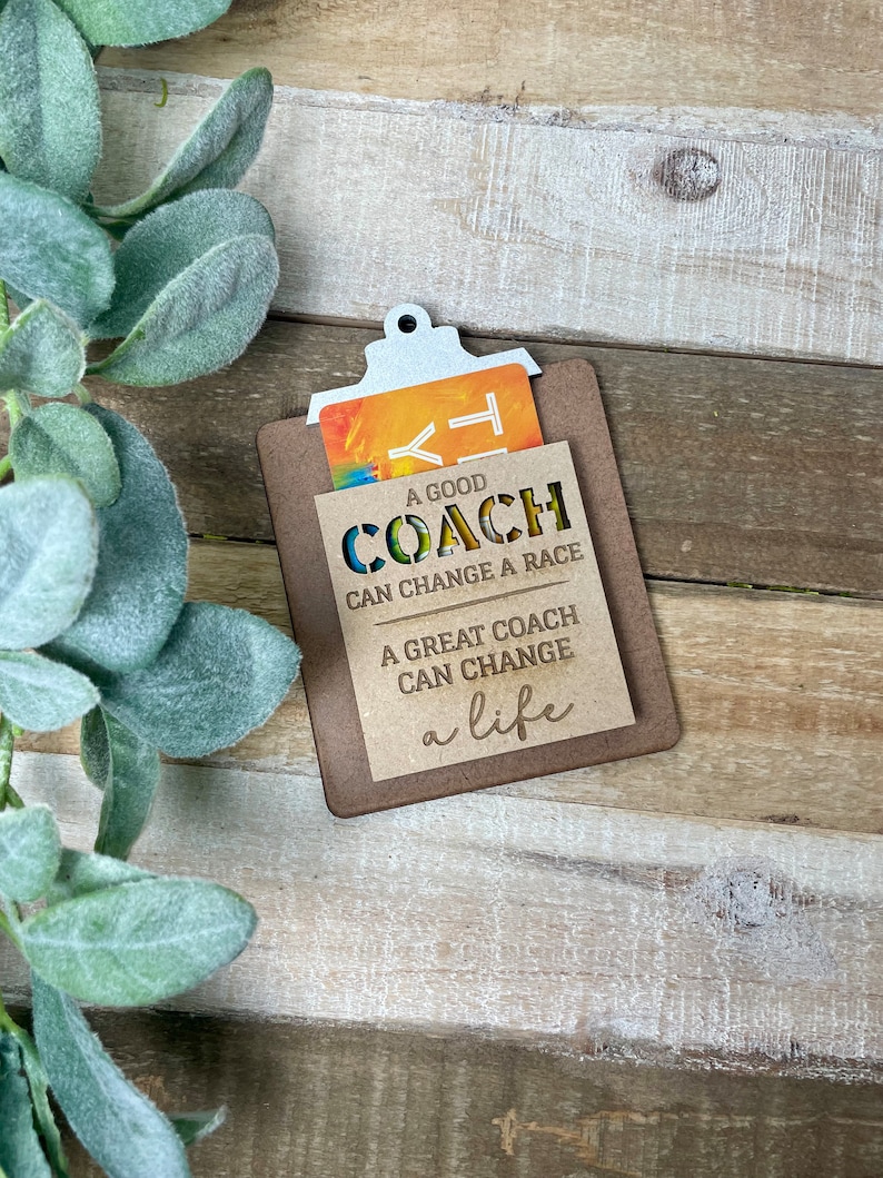 May include: A wooden clipboard with a gift card inside. The clipboard has a cutout design that says "A good coach can change a race. A great coach can change a life."