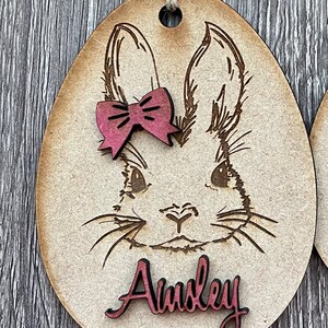 May include: A wooden Easter egg ornament with a laser-cut design of a bunny's face. The bunny has a pink bow and the name "Ainsley" is written in pink on the ornament.