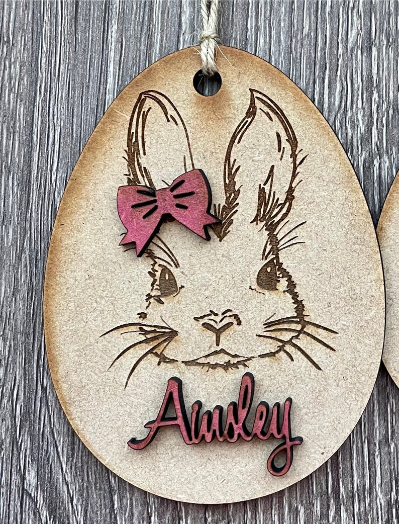 May include: A wooden Easter egg ornament with a laser-cut design of a bunny's face. The bunny has a pink bow and the name "Ainsley" is written in pink on the ornament.