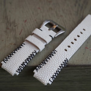 Może przedstawiać: Biały ceramiczny kubek z czarnym uchwytem i napisem "But First, Coffee" pogrubioną czcionką. Kubek jest umieszczony na rozmytym tle, sugerującym przytulne otoczenie.