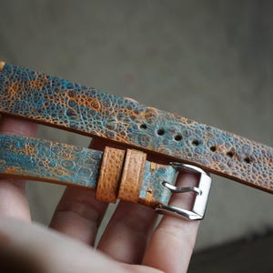 May include: A distressed leather watch strap with a unique blue and orange mottled pattern. The strap features a silver buckle and a contrasting tan leather loop. The watch band is approximately 20 cm long.