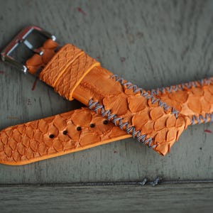 Może przedstawiać: Pomarańczowy pasek do zegarka z teksturowanym wzorem przypominającym łuski. Pasek ma szare szwy i srebrną klamrę. Pasek do zegarka jest wykonany z materiału przypominającego łuski ryb.
