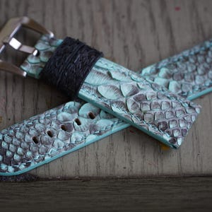 Op de afbeelding: Een horlogebandje met een turquoise en grijs slangenpatroon. De band heeft een zwarte leren lus en een zilverkleurige metalen gesp. De horlogeband ligt op een houten oppervlak.