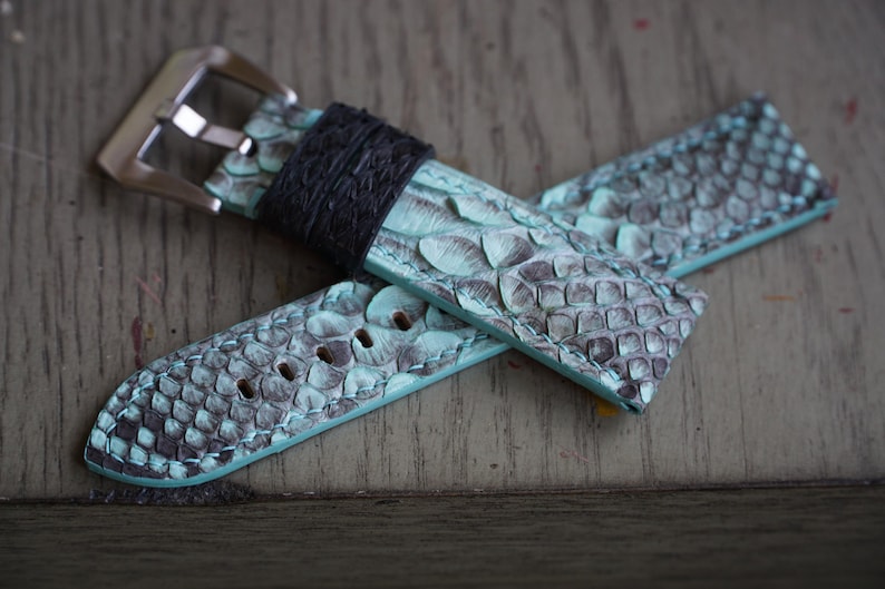 May include: A watch strap with a teal and grey snakeskin pattern. The strap has a black leather loop and a silver-coloured metal buckle. The watch band is lying on a wooden surface.