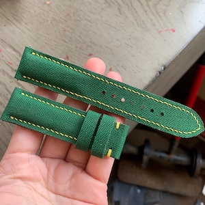 Op de afbeelding: Een groene horlogeband met gele stiksels. De band is gemaakt van een textuurstof en heeft twee lussen en verschillende gaten voor aanpassing. De stiksels zijn zichtbaar langs de randen en rond de lussen.