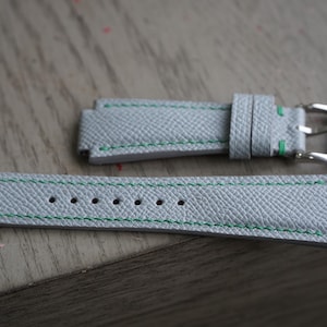 Puede incluir: Correa de reloj gris claro de dos piezas con costuras verdes. La correa tiene una superficie texturizada y una hebilla plateada. La correa del reloj está hecha de cuero y está diseñada para sujetarse a una caja de reloj.