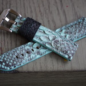 May include: A watch strap made of teal and grey snakeskin-patterned leather. The strap has a silver-coloured buckle and a black leather loop. The watch strap is lying on a wooden surface.