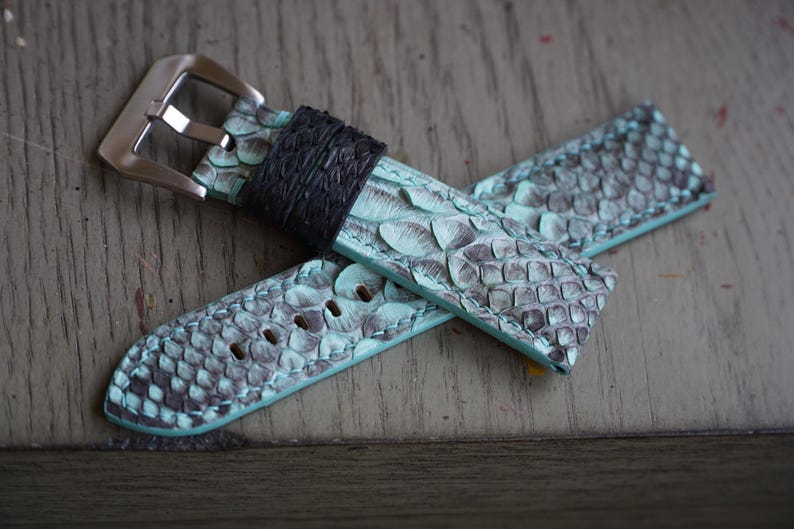 May include: A watch strap made of teal and grey snakeskin-patterned leather. The strap has a silver-coloured buckle and a black leather loop. The watch strap is lying on a wooden surface.