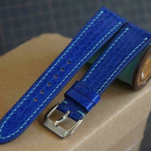 Op de afbeelding: Een blauw leren horlogebandje met lichtblauwe stiksels. Het bandje is te zien op een bruin oppervlak met een spoel draad.