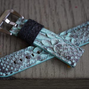May include: A watch strap featuring a teal and grey snakeskin design. It has a black leather keeper and a silver buckle. The strap is made of a textured material and has adjustment holes.