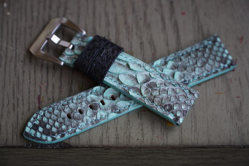 May include: A watch strap featuring a teal and grey snakeskin design. It has a black leather keeper and a silver buckle. The strap is made of a textured material and has adjustment holes.
