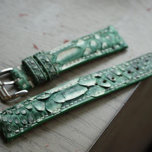 Op de afbeelding: Tweedelig horlogebandje in een groen slangenpatroon. De leren band heeft een zilveren gesp en witte stiksels. De horlogeband heeft een levendige groene kleur met een textuur, schubachtig ontwerp.