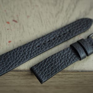 May include: A two-piece black watch strap featuring a textured, pebbled surface finish. The strap includes a series of punched holes for adjustment and a dark metal buckle with two matching leather keepers to secure the end of the watch band.