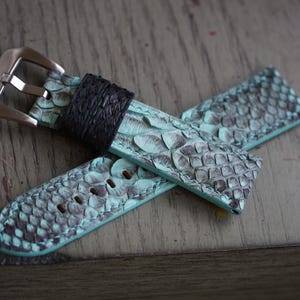 May include: A watch strap with a teal and grey snakeskin pattern. The strap has a black leather band near the buckle and a silver-coloured buckle. The strap is lying on a wooden surface.