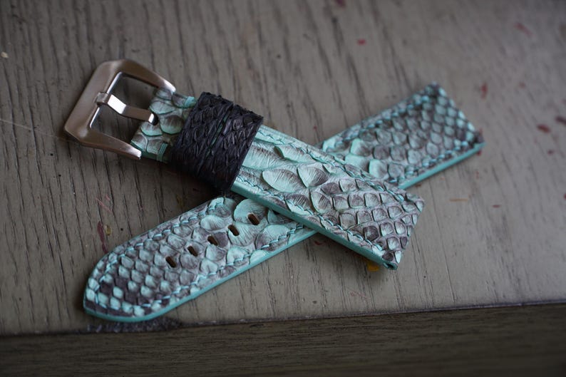May include: A watch strap with a teal and grey snakeskin pattern. The strap has a black leather band near the buckle and a silver-coloured buckle. The strap is lying on a wooden surface.