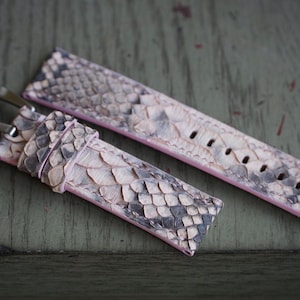 Op de afbeelding: Een horlogebandje van slangenleer in roze, grijze en witte tinten. De band heeft een zilverkleurige gesp en is ontworpen om aan een horloge te worden bevestigd. Het slangenleerpatroon is over de hele band zichtbaar.