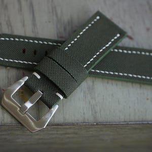 May include: An olive green watch strap with white stitching. The strap has a silver-colored buckle. The watch band is made of a textured material.
