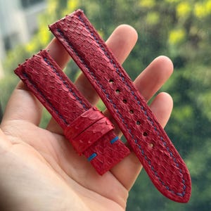 May include: A two-piece red watch strap featuring a textured, scale-like surface pattern. The strap is accented with contrasting blue stitching along the edges and includes two sliding keepers on the shorter piece and buckle holes.