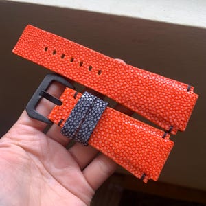 May include: Bright orange watch strap with a textured surface and black stitching. The strap features a black buckle and a section of gray material. The watch band is held in a hand.