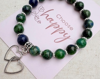 May include: A beaded bracelet featuring round green and dark blue stone beads interspersed with small silver-toned metal spacers. A silver-toned charm consisting of two interlocking open hearts hangs from the bracelet, which rests on a light pink card with the text "Choose happy" printed in a dark script font.