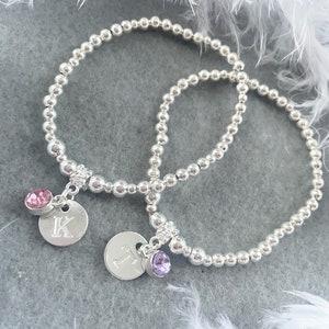 May include: Two silver beaded bracelets with charms. One charm has a pink crystal and the letter "K" engraved on it. The other charm has a purple crystal and the letter "T" engraved on it.