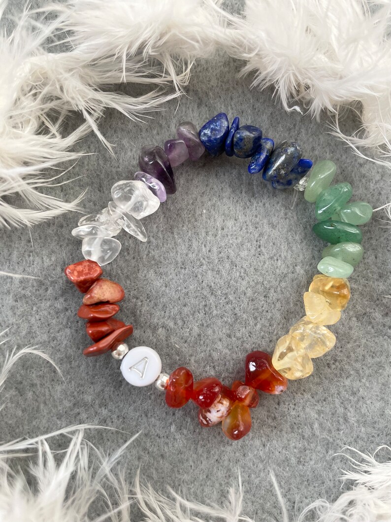 May include: A rainbow chakra bracelet made with natural stones. The bracelet features clear quartz, amethyst, lapis lazuli, green aventurine, citrine, carnelian, and red jasper stones. A silver charm with a triangle symbol is included.