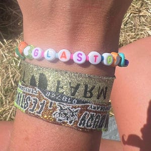 May include: A colorful beaded bracelet with the word "GLASTO" spelled out in white letters. The bracelet is worn on a wrist with two gold glitter wristbands, one with the text "WARWICK" and the other with the text "GLASTO FESTIVAL".