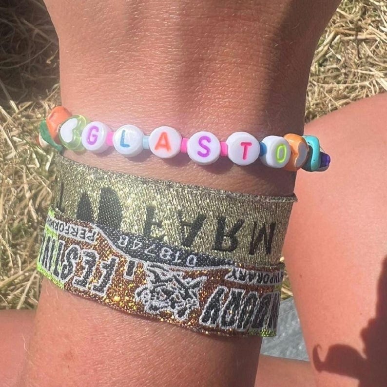 May include: A colorful beaded bracelet with the word "GLASTO" spelled out in white letters. The bracelet is worn on a wrist with two gold glitter wristbands, one with the text "WARWICK" and the other with the text "GLASTO FESTIVAL".