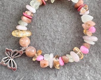 May include: A beaded bracelet featuring a variety of colorful gemstones in shades of pink, yellow, and clear. A silver butterfly charm dangles from the bracelet. The bracelet is displayed on a gray surface.