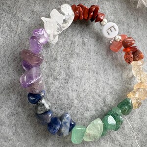 May include: A rainbow chakra bracelet with a silver charm that says "E". The bracelet is made of natural stones in various colors, including red, orange, yellow, green, blue, indigo, and violet.