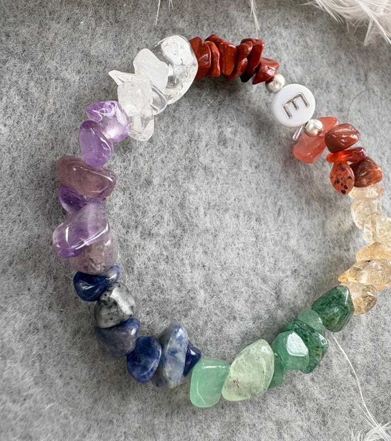 May include: A rainbow chakra bracelet with a silver charm that says "E". The bracelet is made of natural stones in various colors, including red, orange, yellow, green, blue, indigo, and violet.