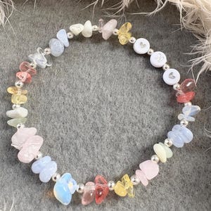 May include: A rainbow gemstone bracelet with silver beads and a charm with the letter "M" and a heart.