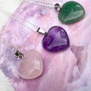 May include: Three heart-shaped gemstone pendants on silver chains. The gemstones are rose quartz, amethyst, and green aventurine. The pendants rest on a purple and white patterned surface.