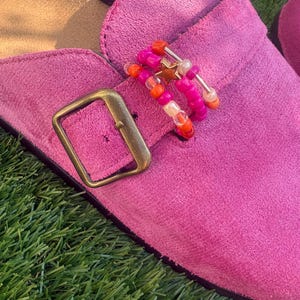 May include: A bright pink suede mule featuring a metallic bronze buckle and a decorative charm accessory attached to the strap. The accessory consists of multiple colourful beads in shades of orange, pink, and white, accented with a small gold star.