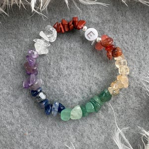 May include: A rainbow crystal bracelet with a silver charm that says "E". The bracelet is made of small, natural stones in shades of red, orange, yellow, green, blue, indigo, and violet.