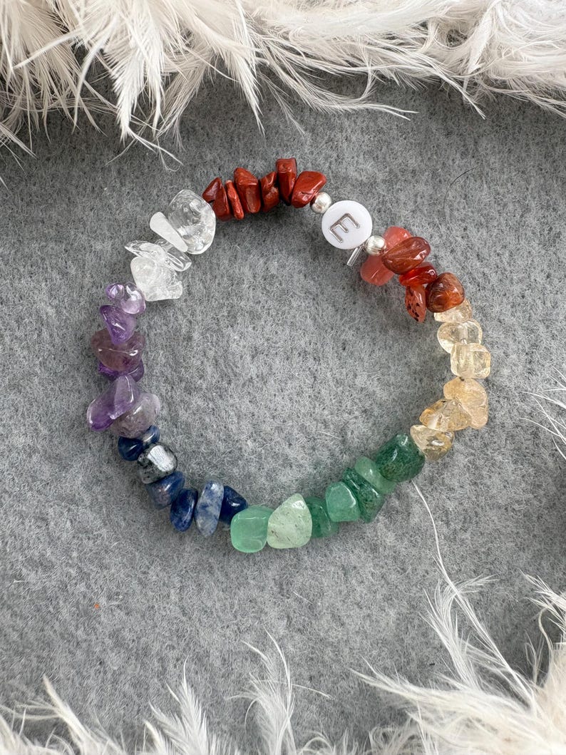 May include: A rainbow crystal bracelet with a silver charm that says "E". The bracelet is made of small, natural stones in shades of red, orange, yellow, green, blue, indigo, and violet.