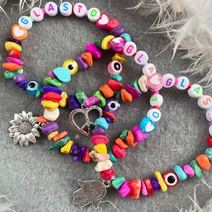 May include: Three colorful beaded bracelets with charms. The bracelets feature colorful beads, letter beads spelling "GLASTO", "GLAS", and "TO", and charms including a sunflower, a peace sign, and a hamsa hand.