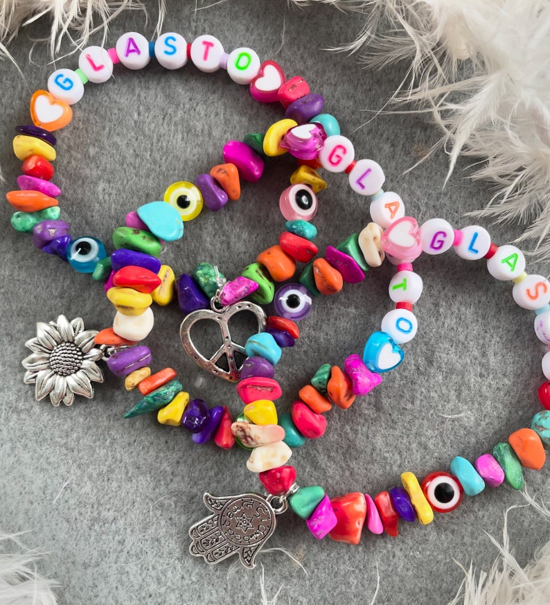 May include: Three colorful beaded bracelets with charms. The bracelets feature colorful beads, letter beads spelling "GLASTO", "GLAS", and "TO", and charms including a sunflower, a peace sign, and a hamsa hand.