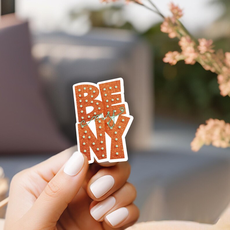 May include: A sticker with the words "BE my NY" in orange letters with white dot lights. The sticker is held by a person with white nail polish.