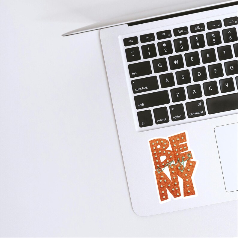 May include: A sticker on a laptop with the words "BE easy NY" in orange and white. The letters are designed to look like marquee lights. The laptop is silver and white.