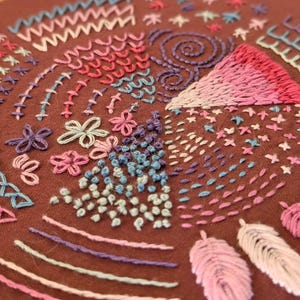 May include: Close-up of a brown fabric with colorful embroidery. The embroidery features various stitches in shades of pink, blue, purple, and white, including flowers, spirals, and geometric patterns. The craftwork showcases a variety of textures and designs.